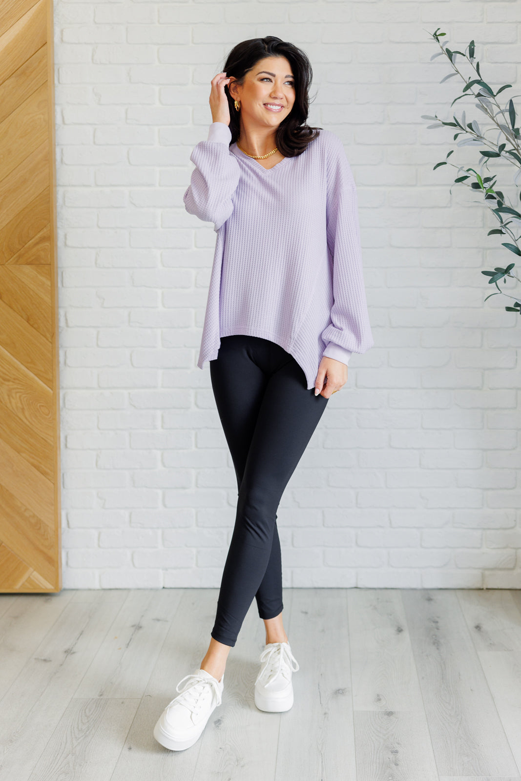 Lavender women’s v-neck top with a relaxed drop shoulder fit, banded sleeve cuffs, scooped asymmetrical hemline, and faux tag detail, styled for an effortless boutique look.