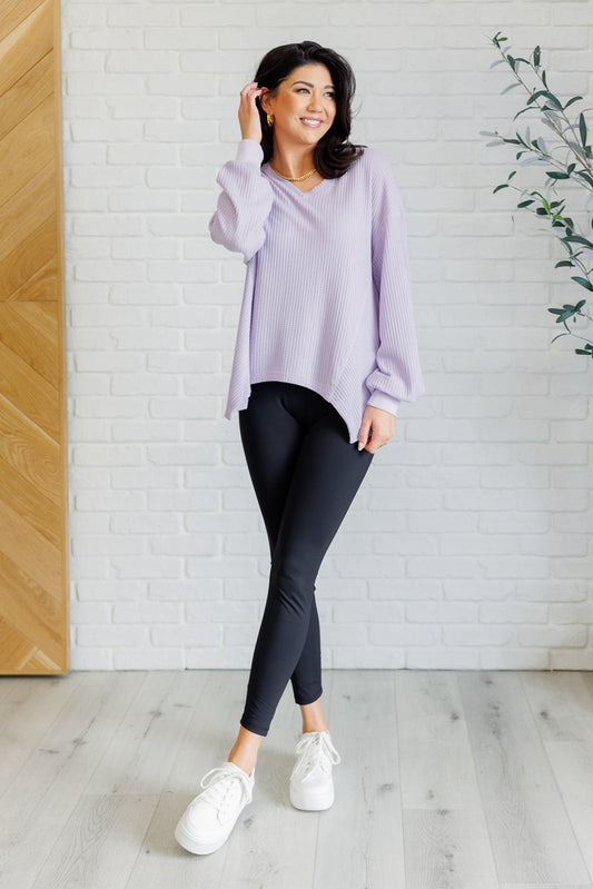 Lavender women’s v-neck top with a relaxed drop shoulder fit, banded sleeve cuffs, scooped asymmetrical hemline, and faux tag detail, styled for an effortless boutique look.