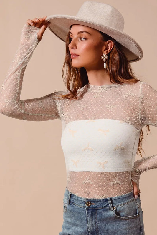 Woman wearing a sheer long-sleeve top with a white base and bow patterns, paired with a wide-brimmed hat and jeans.